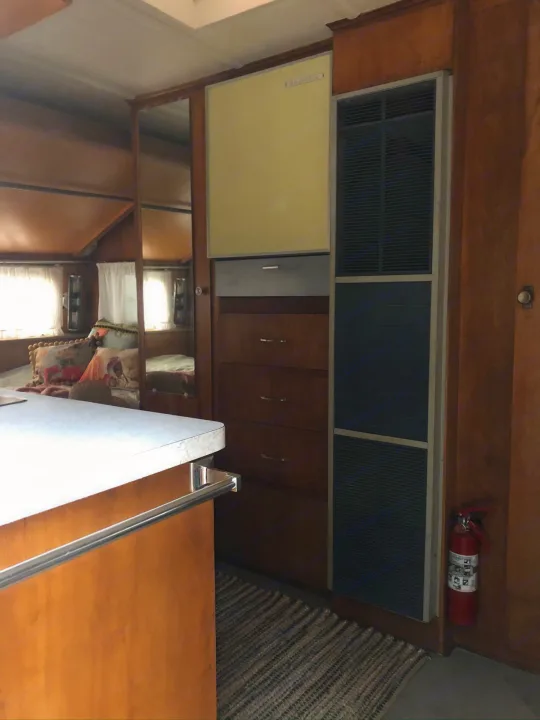 Explore this vintage RV rental interior: warm wood tones, compact fridge, mirrored wall, and fire extinguisher for safe travels.. Holiday Rambler Traveler 1965