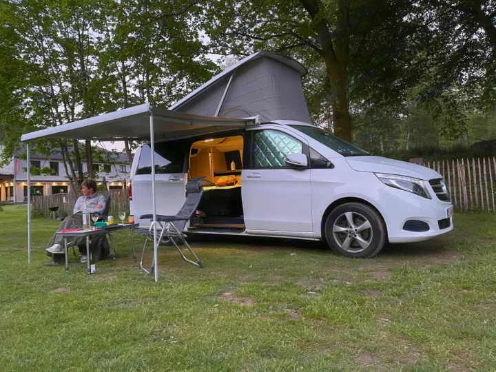 Relaxing with an rv rental camper van at dusk. Pop-top raised, awning out, person enjoying drinks at a campsite.. Mercedes Marco Polo 2022
