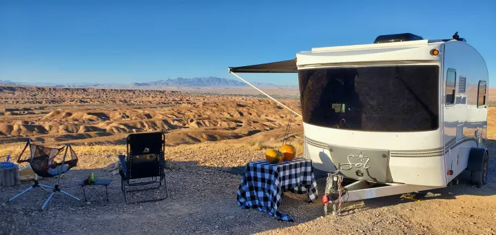 An RV rental parked with an awning extended, two chairs, and a table with pumpkins, overlooking a vast desert landscape and distant town.. InTech RV Sol Horizon 2020