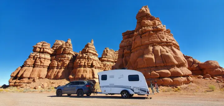 RV rental adventure: Grey SUV towing a white travel trailer with bikes parked near towering desert rock formations under a blue sky.. InTech RV Sol Horizon 2020