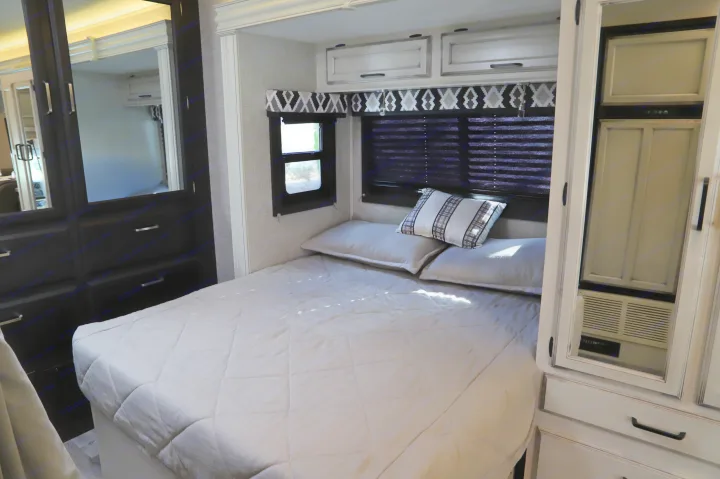 Modern RV rental bedroom interior with a comfy bed, striped pillow, white cabinets, and mirrored wardrobe on the left.. Entegra 24B 2022