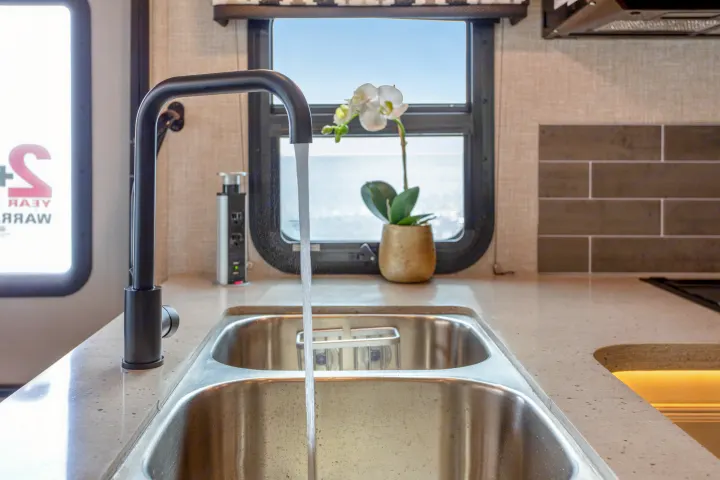 Modern RV rental kitchen with running black faucet over double sink, light countertop, window view, and an orchid.. Other Other 2022