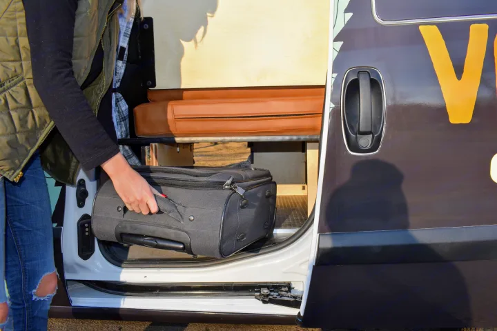 Person loads a black suitcase into an RV rental with a side sliding door, showing its interior wooden panels and brown cushions.. Dodge Promaster City 2021