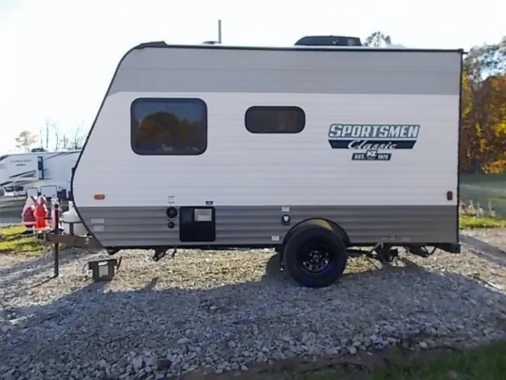 White & grey Sportsmen Classic travel trailer RV rental, parked on gravel. Other RVs and holiday decor in background.. Sportsmen Classic 2022