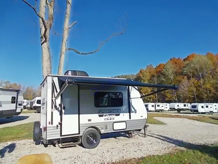 A 'SPORTSMEN Classic' travel trailer with awning extended in an RV rental lot, with other campers and autumn trees under a blue sky.. Sportsmen Classic 2022