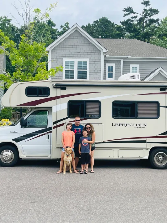 Smiling family with their dog poses in front of a large beige RV rental, ready for a road trip adventure!. Forest River Coachmen Leprechaun 2019