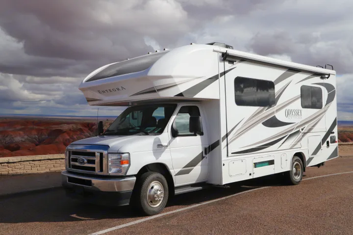 White Entegra Odyssey **RV rental** parked at a scenic overlook, showcasing a vast, colorful desert landscape under cloudy skies.. Entegra Coach Odyssey 2021