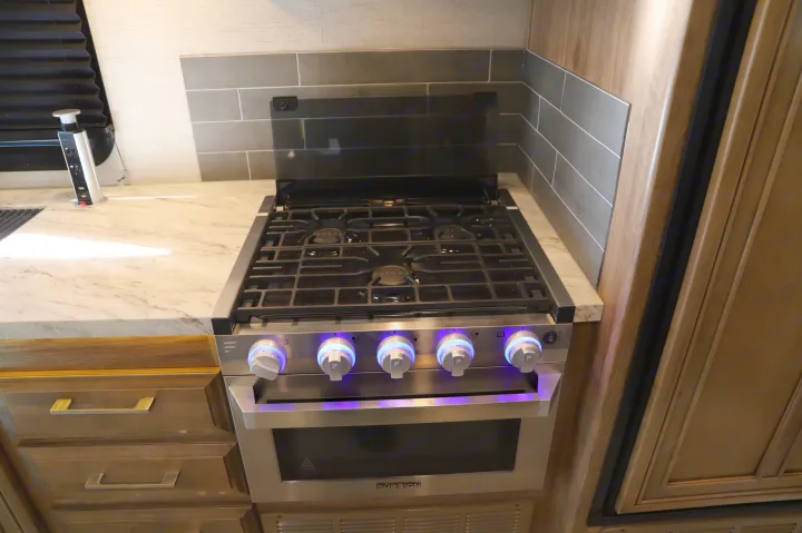 Modern RV rental kitchen with a stainless steel stove/oven featuring illuminated knobs, marble-look countertop, and tile backsplash.. Entegra Coach Odyssey 2021