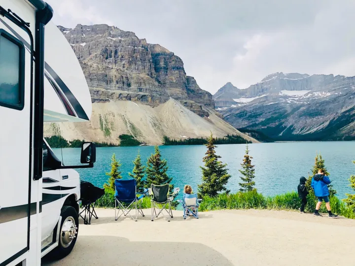 A white RV rental parked by a turquoise lake, with majestic mountains. People relax in chairs & kids explore the scenic view.. Jayco Redhawk 2018