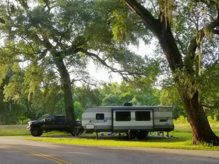 A black pickup truck and 'Sportsmen' RV rental parked under large, mossy trees by a road, perfect for camping.. Sportsmen SE 2021