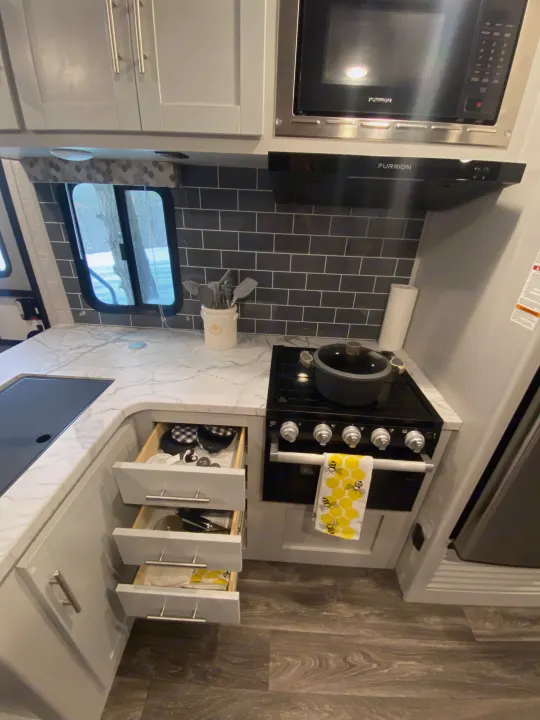 Modern RV rental kitchen featuring a stove, microwave, sink, light cabinets & open drawers. Outside, a snowy treeline.. Keystone RV Bullet Ultra Lite 2021