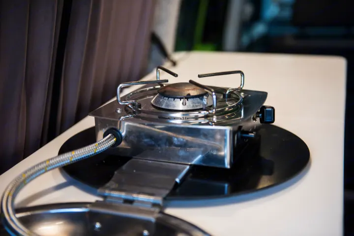 Compact stainless steel gas stove on a white counter, featuring a single burner and connected gas line, perfect for an RV rental.. Volkswagen Caddy Maxi 2017