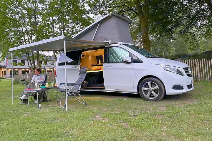 Serene rv rental setup: a white pop-top camper van with awning extended, side door open, and person relaxing outside.. Mercedes Marco Polo 2022
