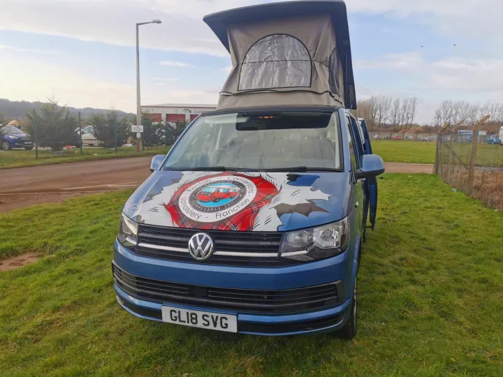 Blue VW pop-top campervan, advertising RV rental & conversions with a tartan decal, parked on grass outdoors.. Volkswagen T5 2018