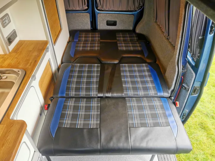 RV rental campervan interior showing a convertible bed with plaid cushions, a kitchen sink, and electrical outlets.. Volkswagen T5 2018