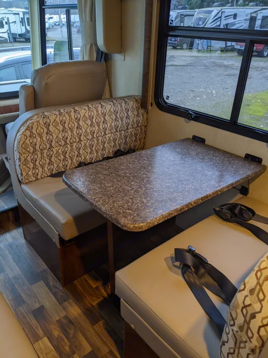 RV rental interior: A functional dinette featuring patterned cushions, a specked table, and safety seatbelts. Other RVs are visible outside.. Coachmen Pursuit 2017