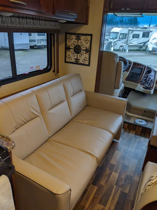 Bright RV rental interior features a long tan sofa, wood cabinetry, cockpit, and window view of other RVs.. Coachmen Pursuit 2017