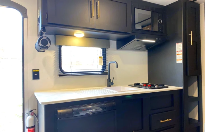 RV rental's compact kitchenette: modern black cabinets, white counter, sink, stovetop, microwave & fridge visible. Ideal for road trips!. Keystone Springdale 1800BH 2022