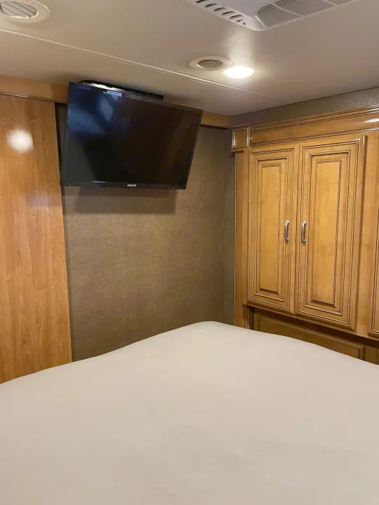 RV rental bedroom showcasing a mounted TV, a bed with light sheets, and wooden storage cabinets.. Thor Palazzo 2019