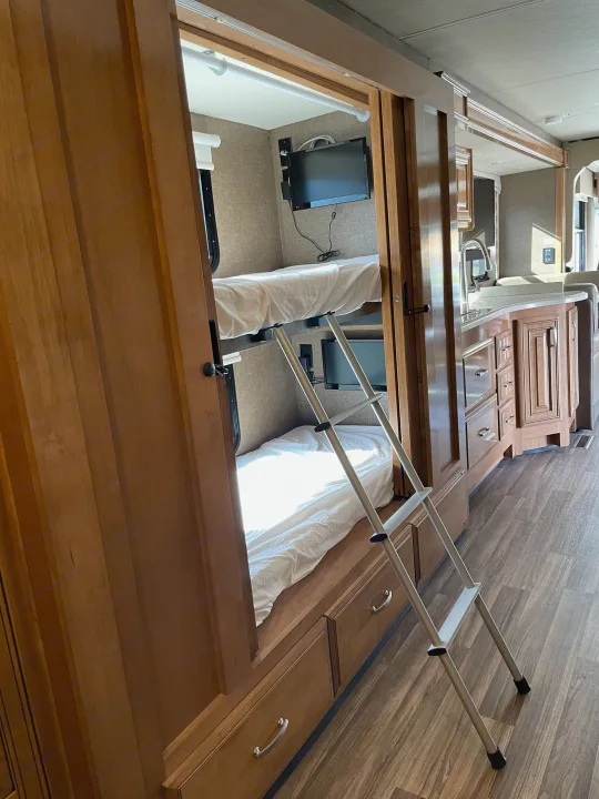 Spacious RV rental bunk beds with TVs, a ladder, and an adjacent kitchen area offer comfort for family travel.. Thor Palazzo 2019