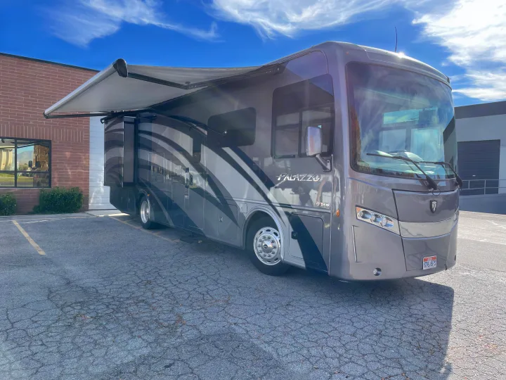 A sleek grey Thor Palazzo RV rental with an extended awning, parked in a lot on a sunny day.. Thor Palazzo 2019