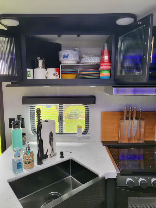 An organized RV rental kitchen featuring a sink, faucet, paper towels, knives, and shelves stocked with dishes and cups.. Forest River Cherokee 2021