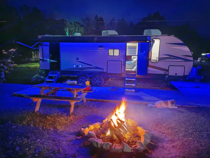 Perfect RV rental evening! Campfire glow, blue-lit travel trailer, and a dog resting at the cozy campsite.. Forest River Cherokee Alpha Wolf 2021