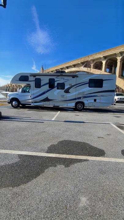 A Freedom Elite Class C RV rental is parked on an asphalt lot, with a large stone arched bridge visible in the background.. Thor Motor Coach Freedom Elite 2015