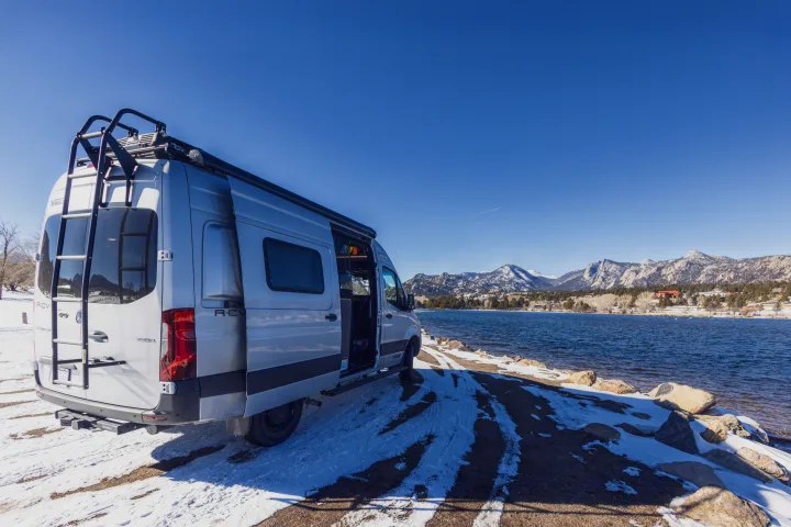 Silver RV rental by a tranquil lake with snow-dusted ground and majestic mountains under a clear blue sky.. Winnebago Revel 2022