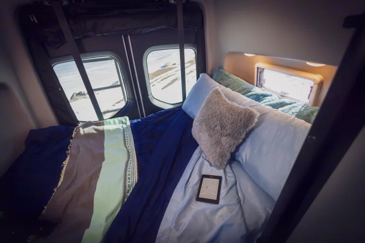 Cozy RV rental bed with a view of a snowy landscape and an e-reader, perfect for a winter escape in a camper van.. Winnebago Revel 2022