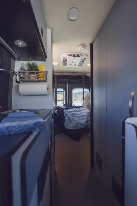 This RV rental interior showcases a compact kitchen area on the left and a cozy bedroom with rear window views, ideal for mobile living.. Winnebago Revel 2022