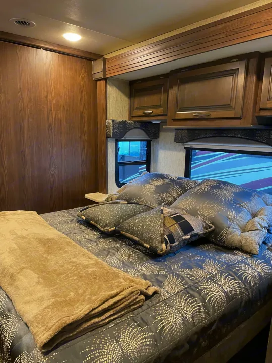 Step inside this cozy RV rental bedroom. Features a comfortable bed, overhead cabinets, and large windows.. Coachmen Mirada 2018
