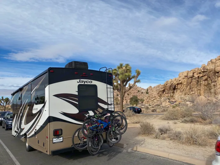 An RV rental (Jayco Greyhawk) with bikes attached, parked in a desert landscape with Joshua trees and rock formations under a blue sky.. Jayco Greyhawk 2018