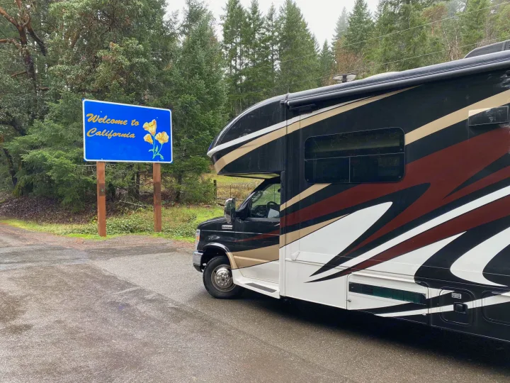 An RV rental pauses at the "Welcome to California" sign, marking a journey through scenic, wet, forested terrain.. Jayco Greyhawk 2018