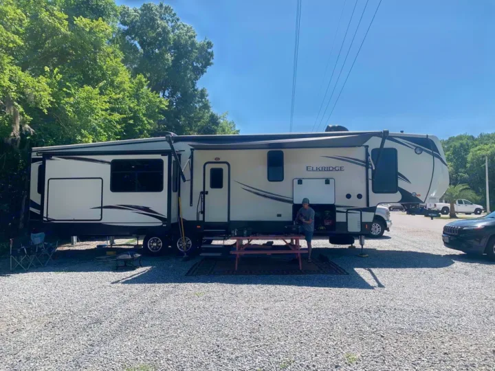 Elkridge RV rental on a sunny gravel campsite, featuring a person, picnic table, and outdoor setup.. Heartland Elkridge 2019
