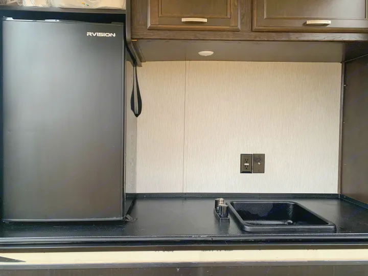 RV rental compact kitchenette featuring a black mini-fridge, sink, and dark countertop with outlets against a textured wall.. Heartland Elkridge 2019
