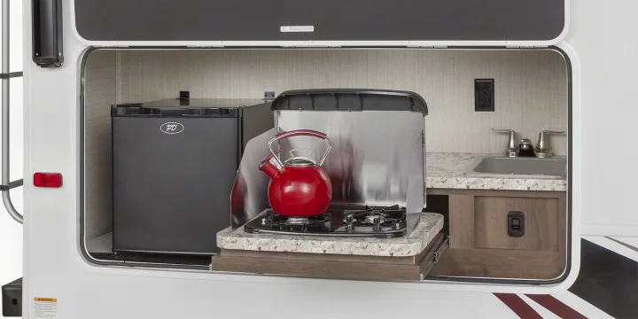 RV rental's outdoor kitchen: compact mini-fridge, sink, and slide-out cooktop with a red kettle.. Jayco Redhawk 2018