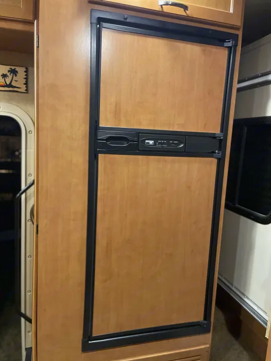 Inside an RV rental: a compact refrigerator with light wood panel door, black trim, and a visible control panel. Essential for travels.. Thor Freedom Elite 2017