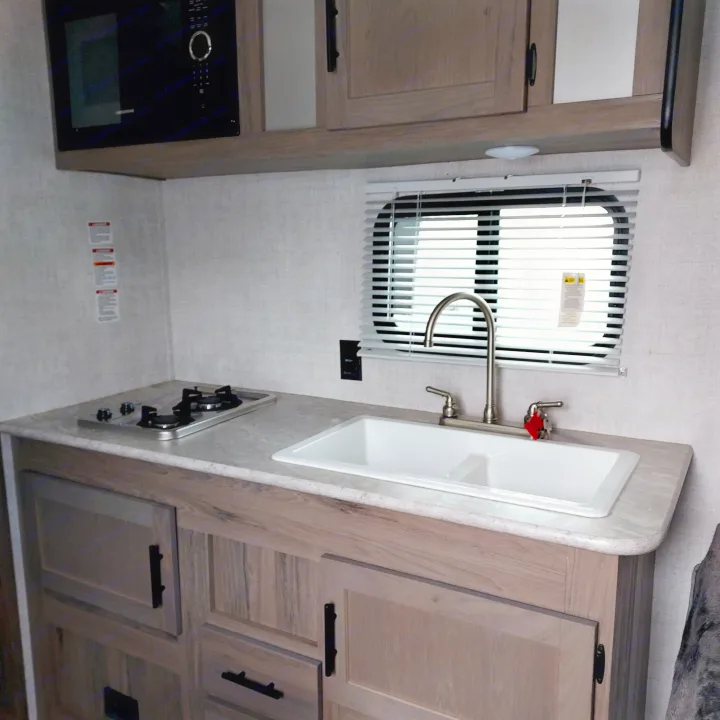RV rental kitchenette with built-in microwave, two-burner stove, and sink. Light wood cabinets define this compact cooking area.. Amerilite 197BH 2021