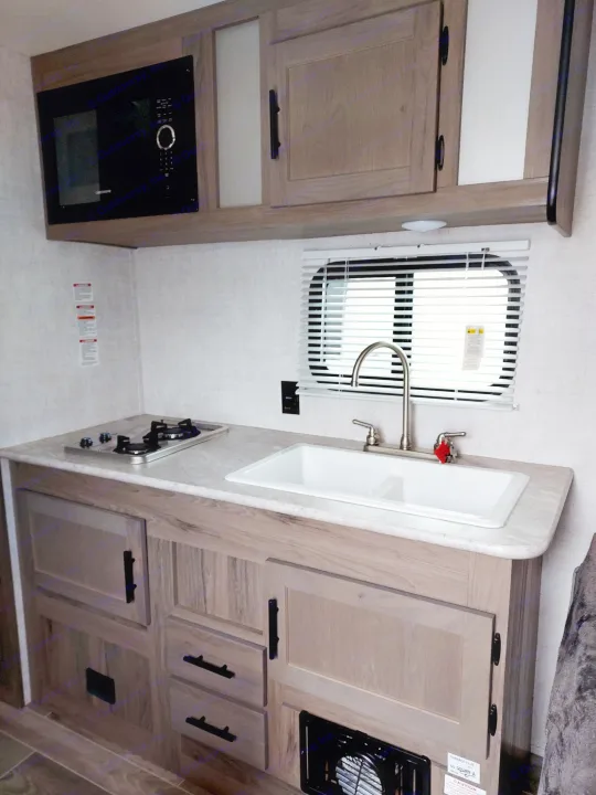 Inside an RV rental: a compact kitchen with a built-in microwave, two-burner stove, sink, and light wood cabinetry.. Amerilite 197BH 2021