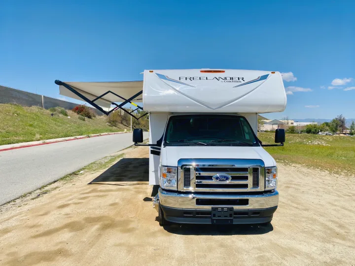 White Class C Freelander RV rental parked on dirt, awning extended, under a blue sky. Ready for outdoor travel.. Coachmen Freelander 2021