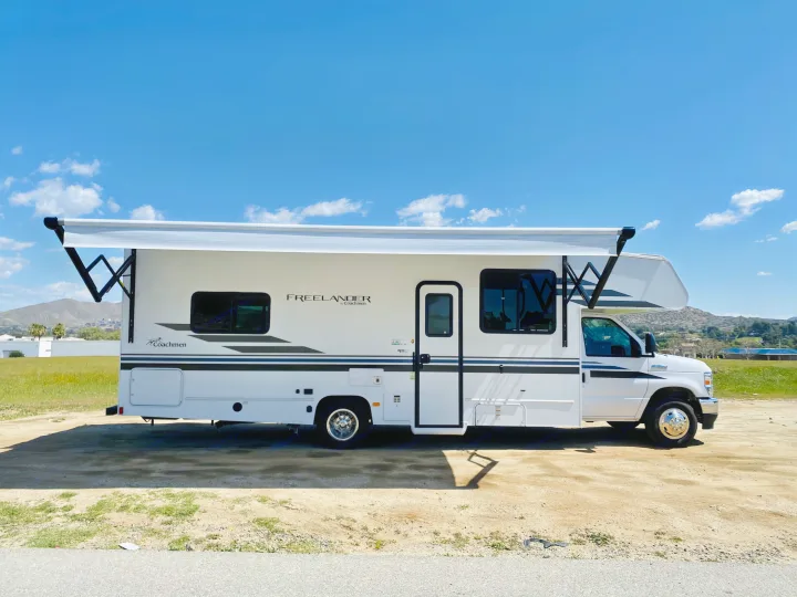White Coachmen Freelander Class C RV rental with awning extended, parked on a dirt lot under blue sky.. Coachmen Freelander 2021