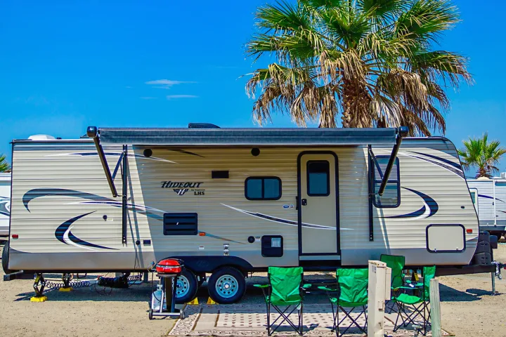 This Hideout travel trailer, perfect for an RV rental, is set up with an awning, grill, and chairs under palm trees.. Keystone Hideout 2018