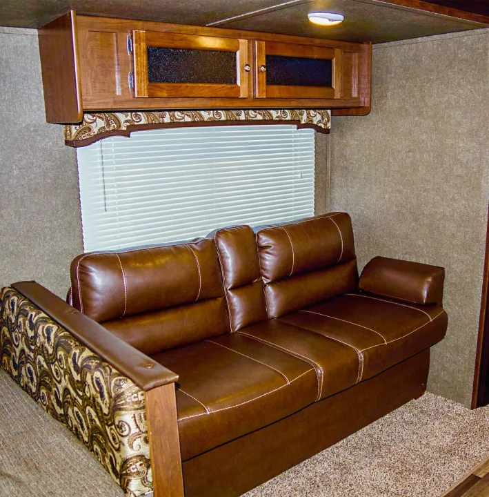 Inside an RV rental: a cozy living area with a brown sofa, patterned armrest and valance, wooden cabinets, and window blinds.. Keystone Hideout 2018
