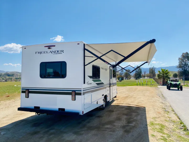 A white Coachmen Freelander RV rental is parked on a sunny dirt lot with its awning extended, ready for outdoor adventures.. Coachmen Freelander 2021
