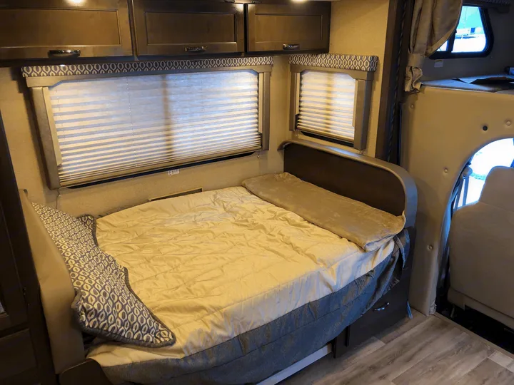 Cozy RV rental interior, featuring a comfortable bed, overhead cabinets, and large windows with horizontal blinds.. Thor Freedom Elite 2018