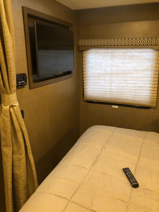 A cozy RV rental bedroom features a comfortable bed with a white comforter, wall-mounted TV, and window with blinds.. Thor Freedom Elite 2018