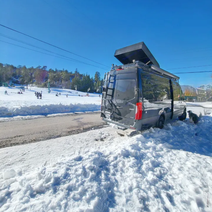 Black camper van (RV rental) parked in snow by a ski slope with people enjoying winter activities. A dog is leashed.. Mercedes-Benz Sprinter 2019