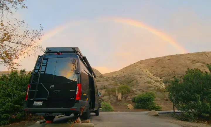 A black camper van sits under a vibrant rainbow in scenic mountains, an ideal RV rental experience.. Mercedes-Benz Sprinter 2019