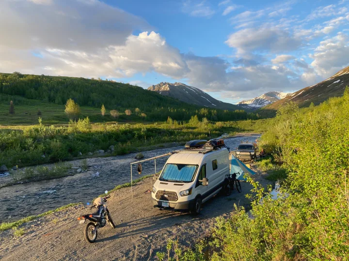 A white RV rental camper van and dirt bike by a scenic river with mountains and lush greenery, perfect for remote camping.. Ford Transit Custom 2015